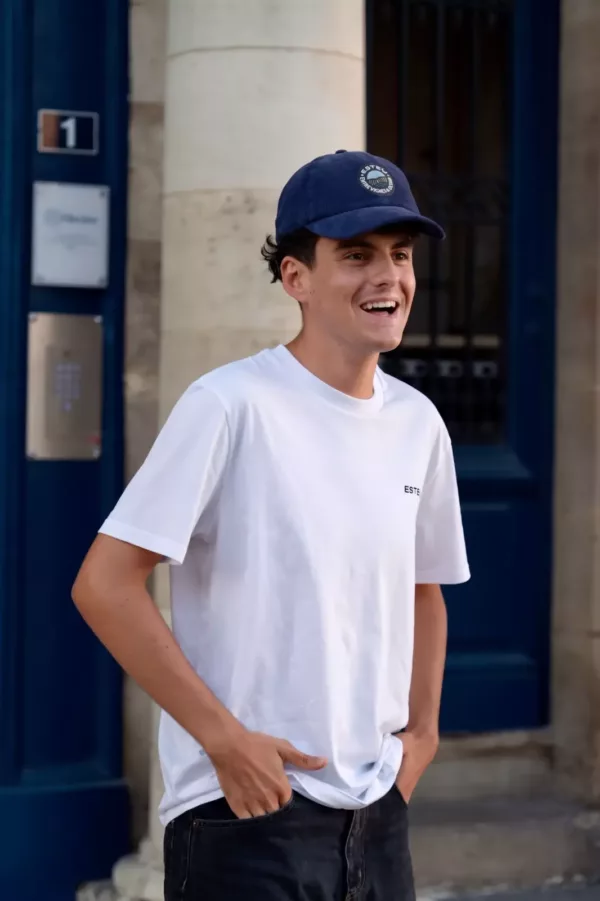 Casquette Le Cap Estey velours côtelé bleu marine portée en ville Homme portant la casquette Le Cap Estey en velours côtelé bleu marine avec un t-shirt blanc, devant une porte ancienne