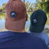 Casquettes Le Cap Estey camel et vert foncé portées à deux Deux casquettes Le Cap Estey en velours côtelé, camel et vert foncé, portées de dos au bord de l’eau
