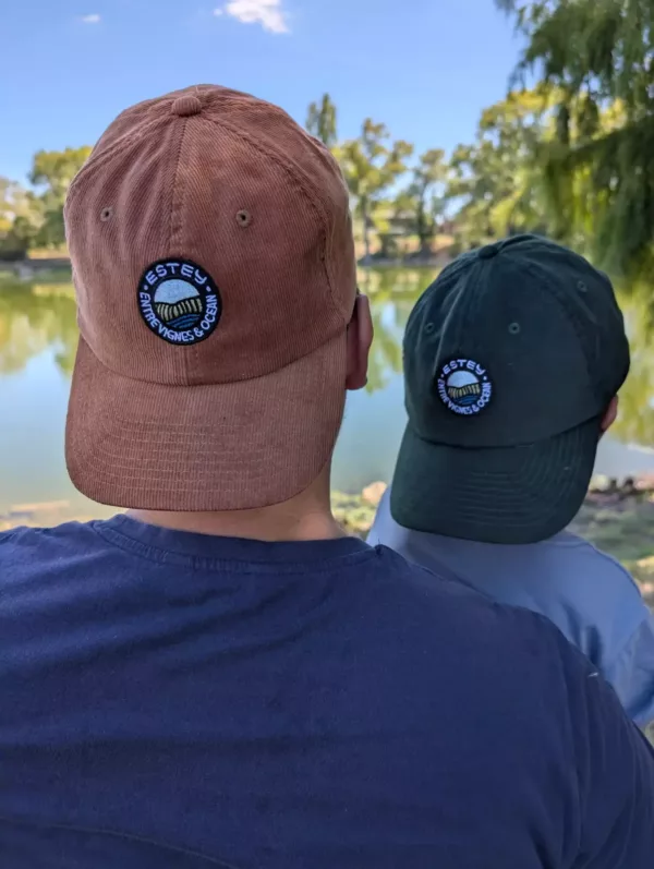 Casquettes Le Cap Estey camel et vert foncé portées à deux Deux casquettes Le Cap Estey en velours côtelé, camel et vert foncé, portées de dos au bord de l’eau
